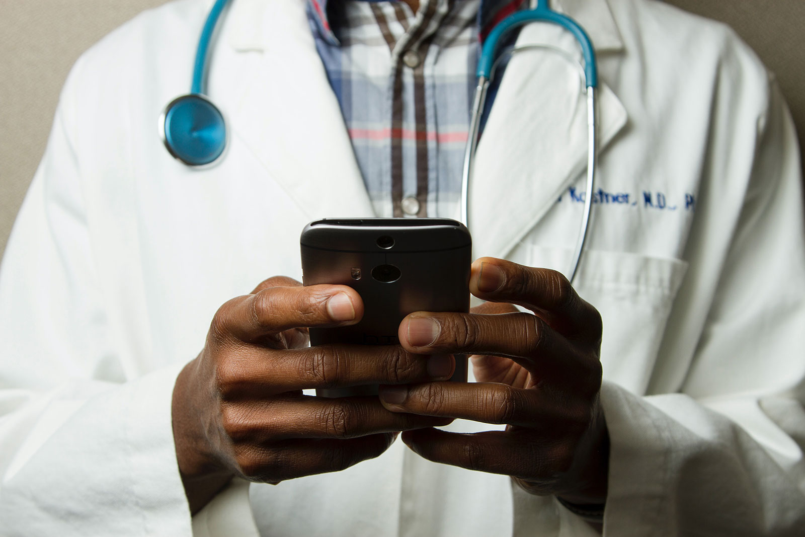 Closeup of the hands of a Black healthcare professional holding a mobile phone and wearing a white coat and stethoscope