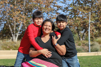 Native American woman and her two boys embracing her while seated on the lawn outdoors with birch trees in the backgroud