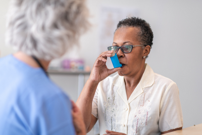 An older black woman uses an inhaler in front of a medical professional