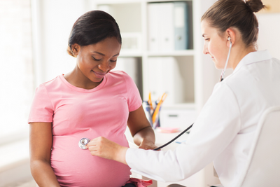 A white female doctor holds a stethoscope to the belly of a pregnant black woman wearing a pink shirt.