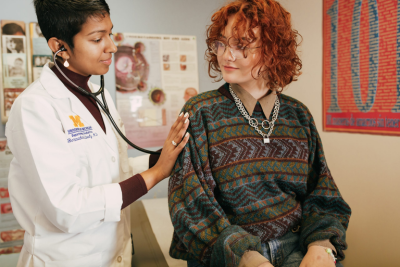 A young female doctor in a white coat attends to a young female patient in a sweater.