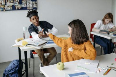young teen passing art supplies to another seated at the neighboring desk